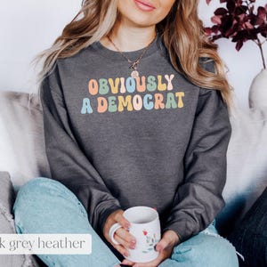 May include: A dark grey heather sweatshirt with the text "OBVIOUSLY A DEMOCRAT" in rainbow colors.