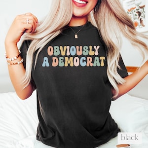 Puede incluir: Camiseta negra con el texto "OBVIOUSLY A DEMOCRAT" en letras de colores de estilo retro.