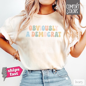Puede incluir: Una camiseta blanca con el texto "OBVIOUSLY A DEMOCRAT" en colores arco&iacute;ris. El texto est&aacute; en un estilo de fuente retro.