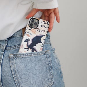 May include: A white phone case with a floral design and a black dragon illustration. The case is in a back pocket of blue jeans.