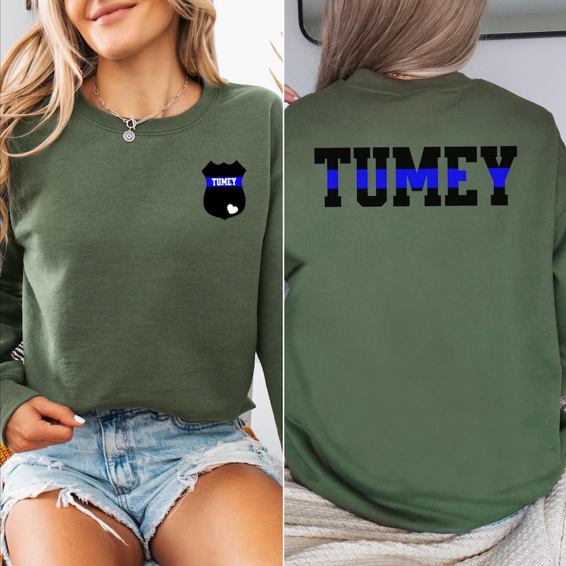 K&ouml;nnte beinhalten: Ein gr&uuml;ner Pullover mit einem schwarzen und blauen Polizeibadge auf der linken Brust und dem Wort "TUMEY" mit einem blauen Streifen auf der R&uuml;ckseite.