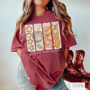 May include: A maroon t-shirt with a graphic of cookie cutters, a rolling pin, a bowl of dough, and a gingerbread man. The text "Just one more batch" is printed below the graphic.