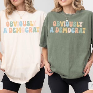 Puede incluir: Dos camisetas con el texto "OBVIOUSLY A DEMOCRAT" en colores arco&iacute;ris. La camiseta de la izquierda es de color marfil y la camiseta de la derecha es de color musgo.