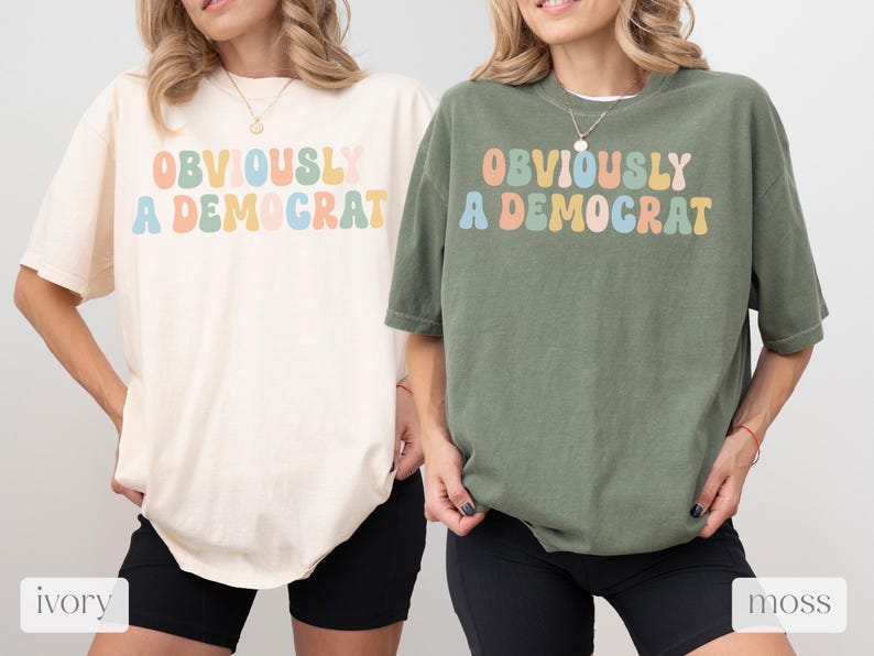 Puede incluir: Dos camisetas con el texto "OBVIOUSLY A DEMOCRAT" en colores arco&iacute;ris. La camiseta de la izquierda es de color marfil y la camiseta de la derecha es de color musgo.