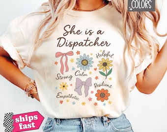 以下が含まれることがあります： 「She is a Dispatcher」の文字と花のグラフィックが特徴のアイボリー色のTシャツ。デザインには、リボンと「Strong」、「Calm」、「Helpful」などの言葉が添えられた花が含まれています。このシャツはComfort Colors素材で作られています。