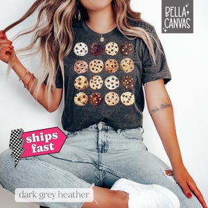 May include: A dark grey heather t-shirt featuring a graphic print of a variety of chocolate chip cookies. The shirt has a crew neck and short sleeves.