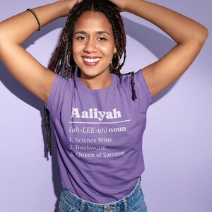 Pu&ograve; includere: Una maglietta viola con il nome "Aaliyah" stampato in bianco, insieme a una definizione e tre tratti: Science Whiz, Bookworm e Queen of Sarcasm. La persona indossa jeans blu.