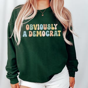 May include: A forest green sweatshirt with the text "OBVIOUSLY A DEMOCRAT" in colorful retro lettering.