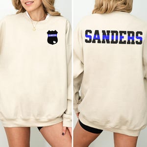 K&ouml;nnte beinhalten: Ein beiger Pullover mit einem schwarzen und blauen Polizeibadge auf der linken Brust und dem Wort "SANDERS" mit einem blauen Streifen auf der R&uuml;ckseite.