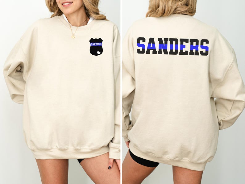 K&ouml;nnte beinhalten: Ein beiger Pullover mit einem schwarzen und blauen Polizeibadge auf der linken Brust und dem Wort "SANDERS" mit einem blauen Streifen auf der R&uuml;ckseite.