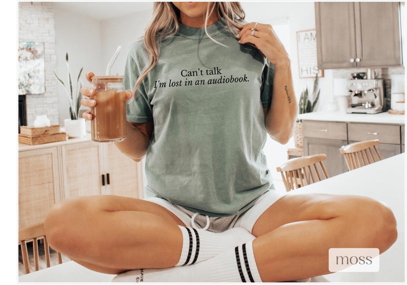 May include: A woman wearing a light green t-shirt with the text "Can't talk I'm lost in an audiobook." The woman is sitting on a white surface and holding a glass with a straw.