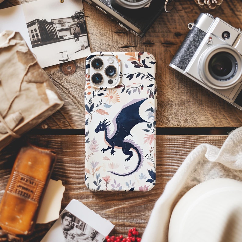 May include: A white phone case with a floral design and a black dragon illustration. The dragon has a blue body and red eyes. The case has a camera lens cutout and a black border.