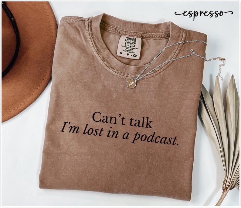 May include: A brown t-shirt with the text "Can't talk I'm lost in a podcast." printed on the front. The shirt is made of a soft, comfortable material and has a relaxed fit.