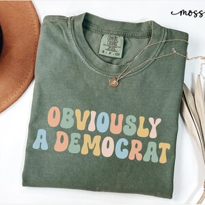 Puede incluir: Una camiseta verde con el texto "OBVIOUSLY A DEMOCRAT" en colores arco&iacute;ris. El texto est&aacute; en un estilo de fuente retro.