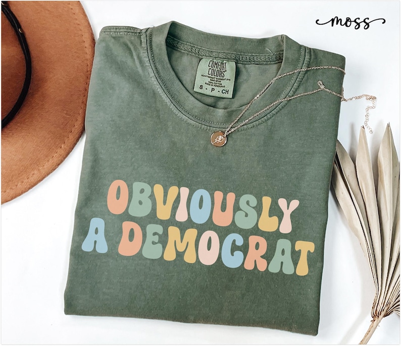 Puede incluir: Una camiseta verde con el texto "OBVIOUSLY A DEMOCRAT" en colores arco&iacute;ris. El texto est&aacute; en un estilo de fuente retro.