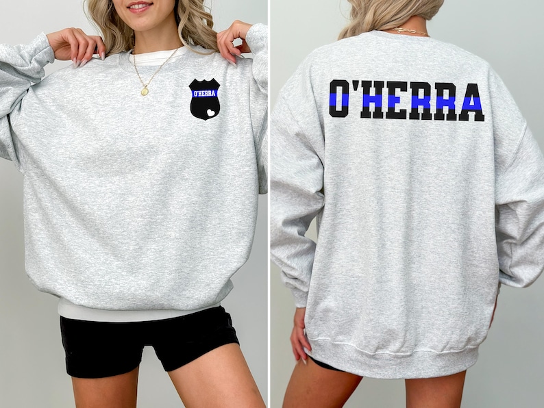 K&ouml;nnte beinhalten: Grauer Sweatshirt mit einem blauen Streifen und einem Abzeichen mit dem Text "O'Herra" auf der Vorderseite. Die R&uuml;ckseite des Sweatshirts tr&auml;gt den Text "O'Herra" mit einem blauen Streifen.