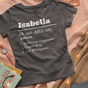 Pu&ograve; includere: T-shirt grigio scuro con il nome "Isabella" in bianco, la sua pronuncia e tre sostantivi: Ingegnere del software, Viaggiatore e Sussurratore di gatti. La maglietta &egrave; esposta su una superficie rosa.