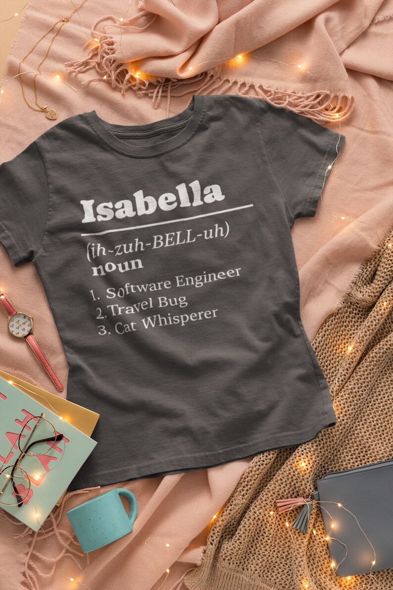 Pu&ograve; includere: T-shirt grigio scuro con il nome "Isabella" in bianco, la sua pronuncia e tre sostantivi: Ingegnere del software, Viaggiatore e Sussurratore di gatti. La maglietta &egrave; esposta su una superficie rosa.