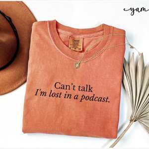 May include: A coral-colored t-shirt with the text "Can't talk I'm lost in a podcast." printed in black.