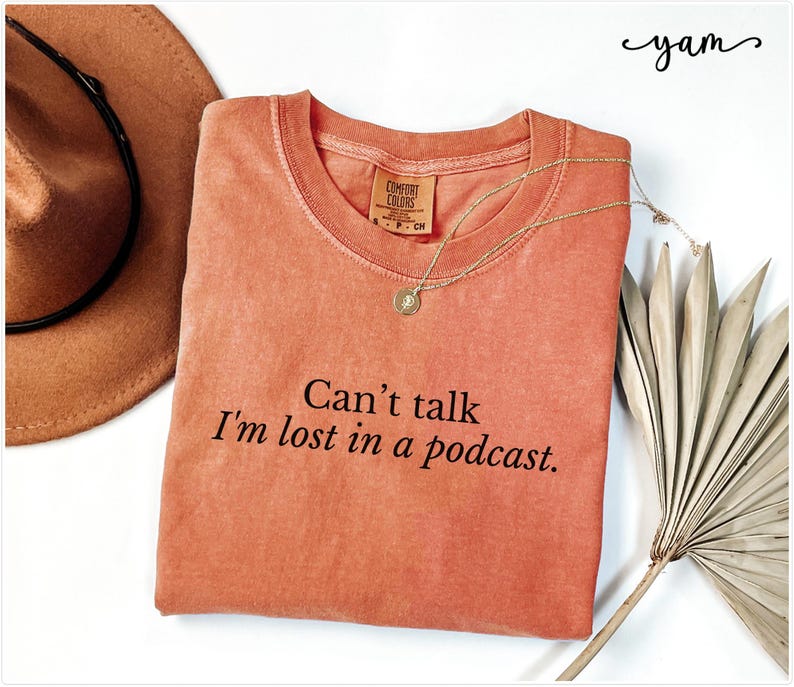 May include: A coral-colored t-shirt with the text "Can't talk I'm lost in a podcast." printed in black.