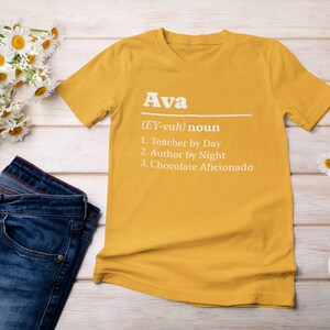 Pu&ograve; includere: T-shirt giallo senape con il nome "Ava" e una definizione stampata in bianco. La definizione include "Insegnante di giorno", "Autrice di notte" e "Appassionata di cioccolato". La maglietta &egrave; abbinata a jeans blu e scarpe da ginnastica bianche.