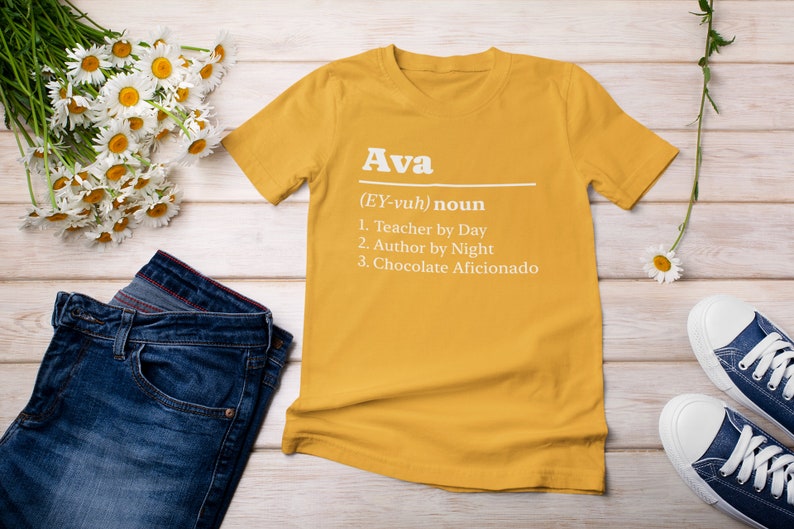 Pu&ograve; includere: T-shirt giallo senape con il nome "Ava" e una definizione stampata in bianco. La definizione include "Insegnante di giorno", "Autrice di notte" e "Appassionata di cioccolato". La maglietta &egrave; abbinata a jeans blu e scarpe da ginnastica bianche.