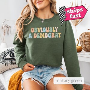 May include: A green sweatshirt with the text "OBVIOUSLY A DEMOCRAT" in rainbow colors. The sweatshirt is a cropped style and has long sleeves. The model is wearing the sweatshirt with denim shorts.