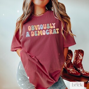 Puede incluir: Una camiseta color granate con el texto "OBVIOUSLY A DEMOCRAT" en letras de colores arco&iacute;ris.