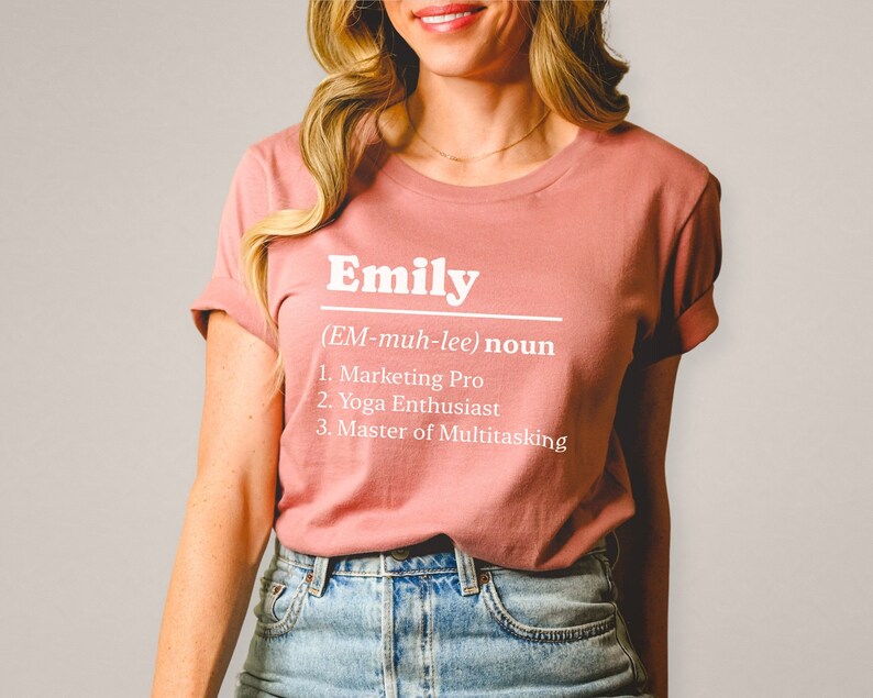 Pu&ograve; includere: T-shirt rosa antico con il nome "Emily" stampato in bianco, accompagnato da una definizione che elenca "Marketing Pro", "Yoga Enthusiast" e "Master of Multitasking". Le maniche sono arrotolate.