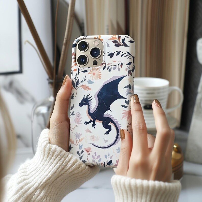 May include: A white iPhone case with a floral pattern and a black dragon design. The dragon has blue wings and is flying in the air. The case has a camera lens cutout.