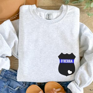 K&ouml;nnte beinhalten: Ein graues Sweatshirt mit einem schwarzen und blauen Polizeibadge-Design. Das Abzeichen hat ein blaues Band mit dem Text "O'Herra" darauf.