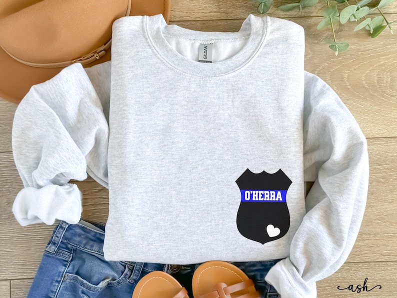 K&ouml;nnte beinhalten: Ein graues Sweatshirt mit einem schwarzen und blauen Polizeibadge-Design. Das Abzeichen hat ein blaues Band mit dem Text "O'Herra" darauf.
