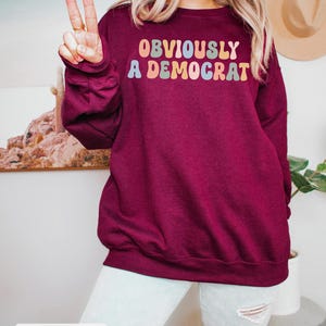 May include: A maroon sweatshirt with the text "OBVIOUSLY A DEMOCRAT" in rainbow-colored letters.