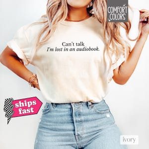 May include: A white t-shirt with the text "Can't talk I'm lost in an audiobook." printed on it. The shirt is being worn by a person with blonde hair and is paired with blue jeans.