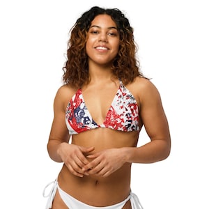 May include: A white bikini set. The triangle top features a red, white, and blue abstract pattern. The bottoms are white with tie closures on each side. The model is smiling and looking at the camera.