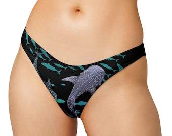 Puede incluir: Braguita de bikini negra con un estampado de tiburón ballena y peces turquesa. El traje de baño presenta un corte de cintura baja y una ilustración detallada de un tiburón ballena con manchas blancas. El fondo es negro con peces turquesa.