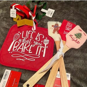 May include: Three red and pink silicone spatulas with wooden handles and Christmas-themed designs. The spatulas are tied together with a white ribbon. A red quilted hot pad with the text "Life is what you bake it" is in the background.