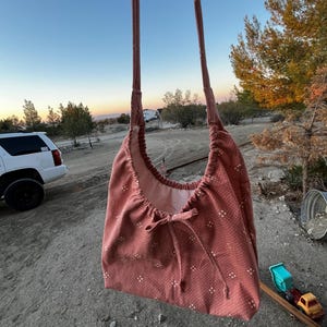 May include: A dusty rose shoulder bag with a drawstring closure and a repeating white dot pattern. The bag has long straps and a light pink interior. The bag is hanging outdoors with a blurred background of a vehicle, trees, and a sunset.