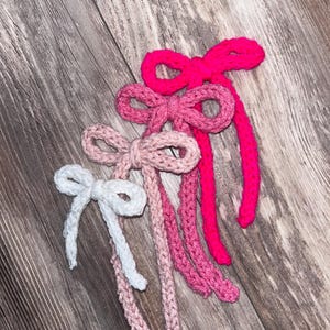 May include: Four crocheted bows in pink, white, and hot pink colors. The bows are tied together and are arranged in a graduated size order.