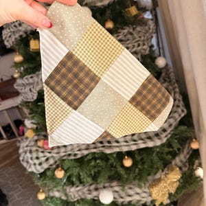 May include: A patchwork Christmas tree skirt in shades of beige, brown, and gold. The skirt is made of various fabric patterns, including polka dots, stripes, and plaid. The skirt is being held up in front of a decorated Christmas tree.