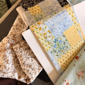 May include: Assortment of fabric squares in various floral and patterned designs. The fabrics include a cream-colored material with brown flowers, a blue and yellow floral patchwork, and a black paisley print. The fabrics are stacked on a white surface.