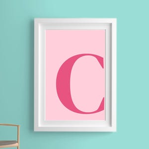 May include: A white framed print with a pink background and a large, bold, pink letter C in the center.