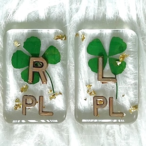May include: Two clear resin squares with gold flecks and a four-leaf clover pressed inside. The squares have the letters "PL" and "R" and "L" printed in gold.