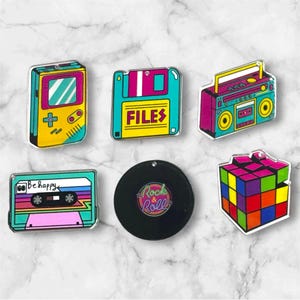 May include: A collection of retro-themed acrylic pins. Includes a yellow and teal handheld game console, a teal floppy disk with "FILES" text, a pink and teal boombox, a cassette tape with "Be happy" text, a black vinyl record with "Rock & Roll" text, and a Rubik's Cube.