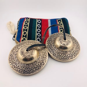 May include: Two small, ornate, brass cymbals with a leather strap. The cymbals have a floral design and are resting on a colorful fabric pouch.