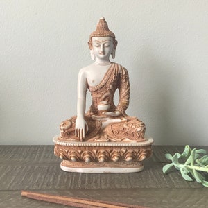 May include: A seated Buddha statue in cream and brown tones. The figure is adorned with detailed patterns on the robe and base, holding a small bowl. The statue is a decorative item, likely for home decor or spiritual practice.