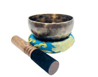 May include: A bronze singing bowl with a decorative design on the bottom, resting on a blue and gold cushion. A wooden striker with a black felt tip is included.