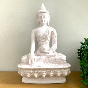 May include: White ceramic Buddha statue in a seated meditation pose. The statue has intricate details on the clothing and base.