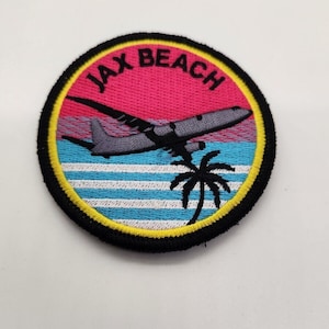Puede incluir: Un parche redondo bordado en negro con un borde amarillo. El parche presenta una puesta de sol rosa, una palmera y un avión volando sobre un océano azul con rayas blancas. El texto "JAX BEACH" está bordado en negro en el parche.