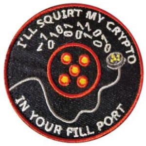 May include: A black and red circular patch with white text that reads "I'll Squirt My Crypto Into Your Fill Port". The patch features a stylized image of a computer plug with a red cable and five yellow lights.